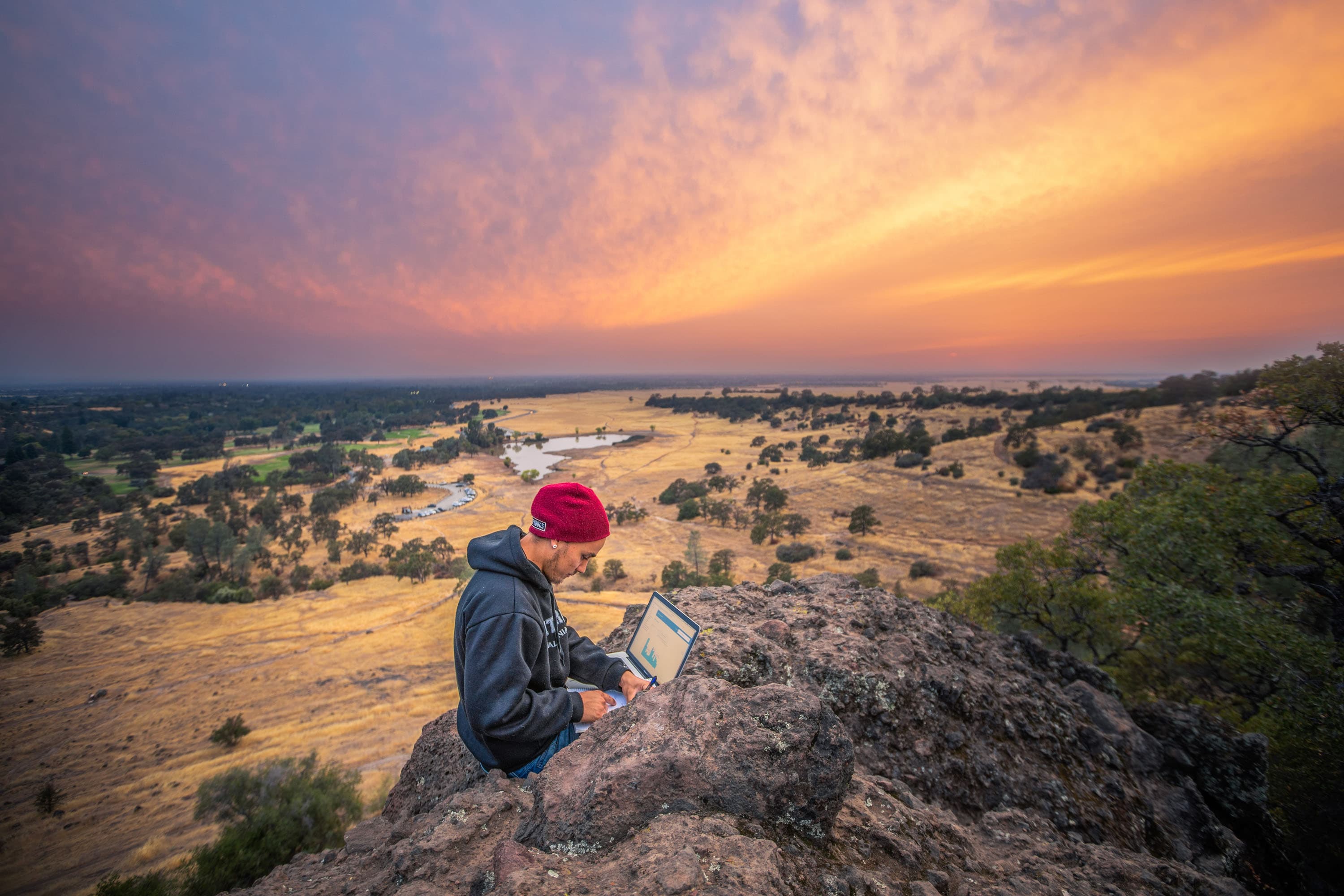 banner image- man on laptop ontop of a mountain overlooking openland and sunset