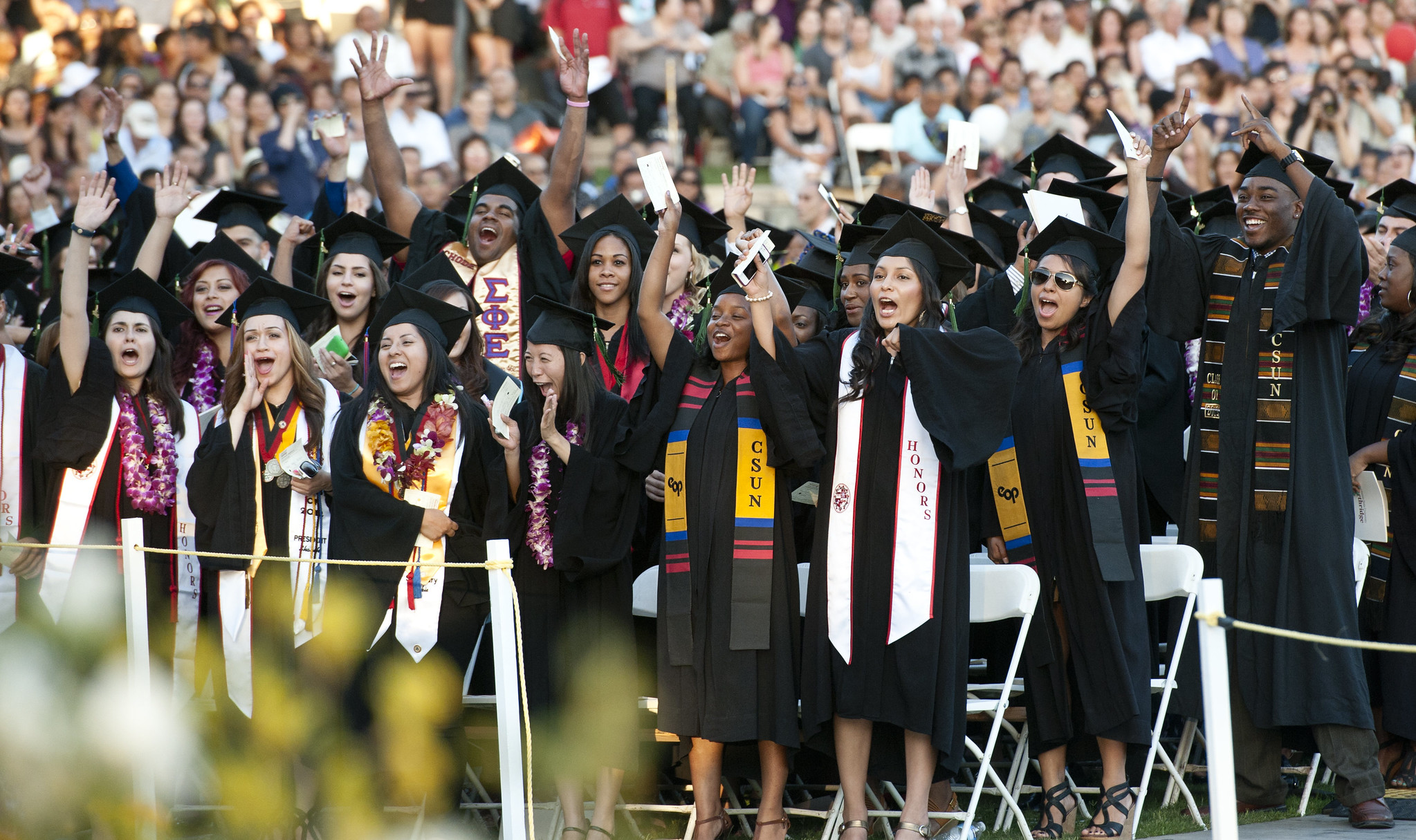 A large group of people wearing graduation caps and gowns cheering and smiling.
