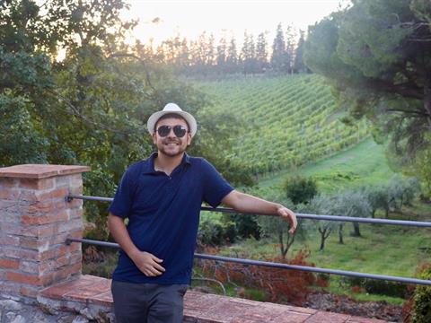 A young person posing in front of rows of grapes growing at a vineyard