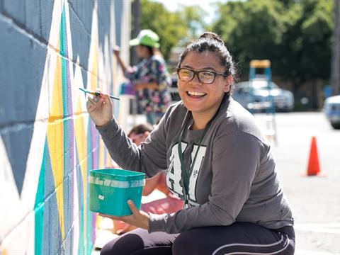 woman with glasses holding a paintbrush and smiling as she squats next to an outdoor wall mural she is painting