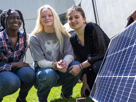 three women sit next to a solar energy panel outdoors