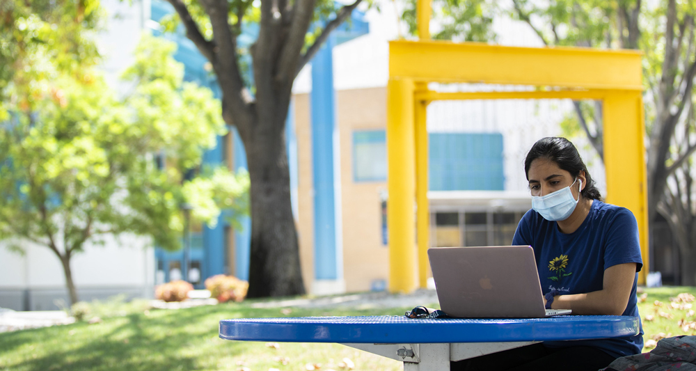 female student wearing a facemask while sitting at an outdoor table using a laptop computer