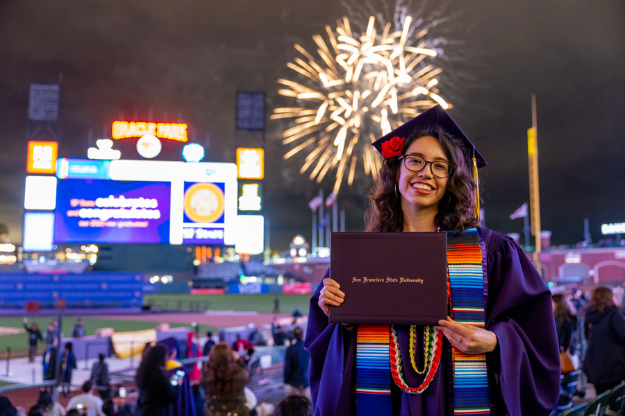 graduating student holding diplomas up with fireworks behind her